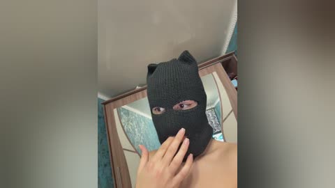 A video of a person wearing a black cat mask, with a hand gently touching the mask's nose, standing in front of a mirror and a turquoise wall.