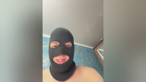 Video of a woman with light skin, wearing a black ski mask with two eyeholes and pink lipstick, standing on a blue carpeted floor, near a window.