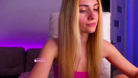 A video of a young Caucasian woman with long, straight blonde hair, wearing a pink strapless top, sitting on a white couch under purple lighting in a modern living room.