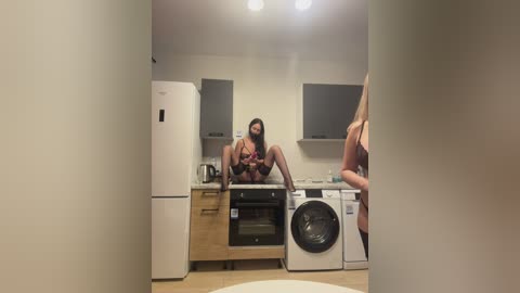 Video of a woman with fair skin, long dark hair, and medium breasts, sitting on a kitchen countertop in black lingerie, legs spread, holding a small bottle. The kitchen has light wooden cabinets, a white fridge, and a washing machine.