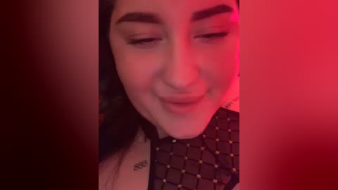 A close-up video of a smiling woman with medium-length dark hair, wearing a black, patterned top, set against a red-lit background.