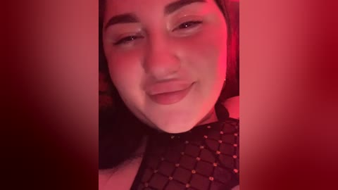 Video of a young woman with light skin and dark hair, wearing a black fishnet top, smiling with a slight pout. The image is bathed in red lighting, creating a sensual, intimate atmosphere.