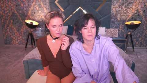 Video of two women in a modern, patterned room. One woman in a brown bodysuit, another in a purple shirt, sitting on a teal sofa.
