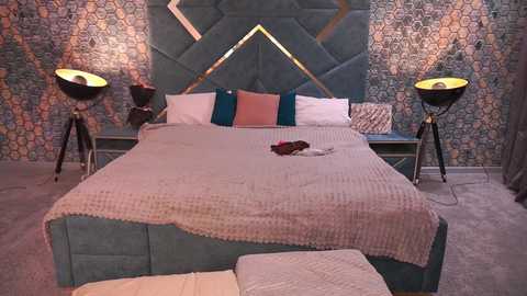Video of a modern bedroom with a teal, tufted headboard, patterned wallpaper, a beige knitted throw on the bed, two black floor lamps, and a plush gray carpet.