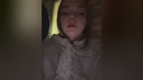 A video of a young woman with fair skin and long, brown hair, wearing a fluffy white robe, seated in a dimly lit room with a brick wall behind her.