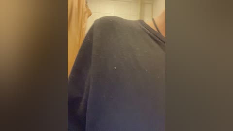 Video of a person in a dark, long-sleeved shirt, likely in a bathroom with a beige tiled wall and a brown shower curtain to the left. The focus is on the person's back.
