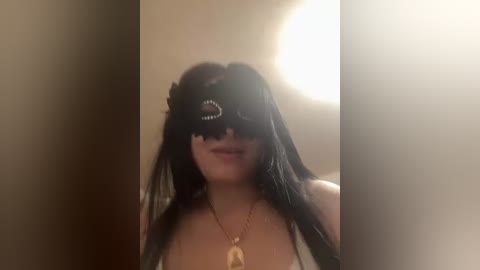 Video of a woman with long black hair, wearing a black mask with a circular design, a gold necklace, and a white top. Background is dimly lit, possibly indoors.
