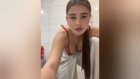 Video of a young woman with long brown hair, wearing a red bra and white towel, leaning forward in a tiled bathroom.