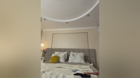 Video of a modern bedroom with beige walls, a tufted gray headboard, white floral-patterned pillows, and a mustard-yellow throw pillow. A yellow lamp and a vase of pink flowers sit on a nightstand. A person, partially visible, lies on the bed.