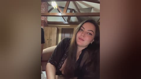 Video of a young, fair-skinned woman with long brown hair, wearing a black sheer robe, leaning against a wooden railing in a rustic, cozy attic room with exposed beams and brick accents.