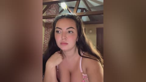 Video of a young woman with long, dark hair, light skin, and full lips, wearing a pink bra, standing in a rustic, wooden-beamed room with exposed brick walls.