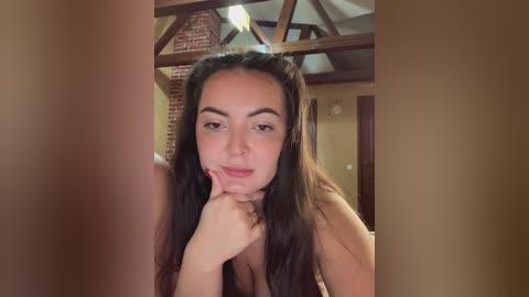 Video of a young woman with long dark hair, light skin, and a delicate facial structure, resting her chin on her hand, set against rustic wooden beams and a brick wall.