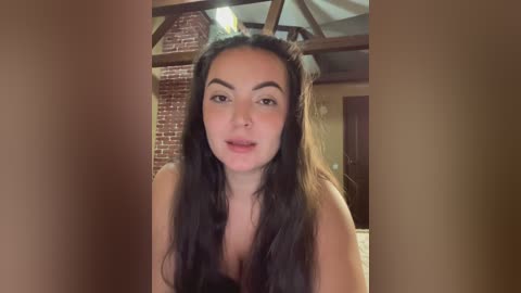 A video of a young woman with long, straight black hair and a light brown complexion, wearing a black top, standing in a rustic, brick-walled room with exposed wooden beams.