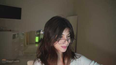 Video of an Asian woman with long, dark hair, wearing glasses, and a white shirt, smiling softly. Background features a kitchen with a microwave, fridge, and dim lighting.