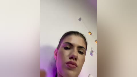 Video of a young woman with light olive skin and dark hair pulled back, looking serious. She stands against a white wall with string lights, creating a soft, warm glow.