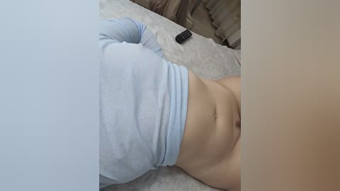Video of a light-skinned person lying on a bed, wearing a light blue long-sleeve shirt, revealing a flat stomach and a black remote control nearby.