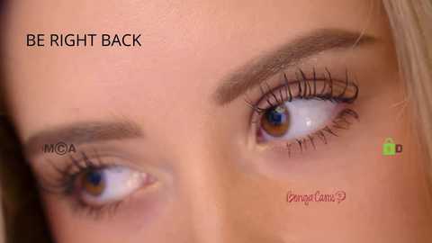 A close-up video of a woman's eye, with the text \"BE RIGHT BACK\" in black, and a green \"MGA\" logo on her cheek. The eye has thick, dark eyebrows, long eyelashes, and light brown skin.