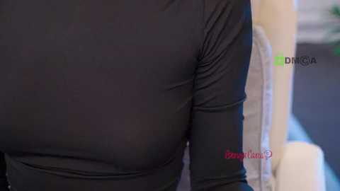 A close-up video of a person wearing a tight-fitting, black long-sleeve shirt with a smooth texture, revealing their prominent, round breasts. The background is blurred, featuring a beige chair and green and black text reading \"PornHub.\
