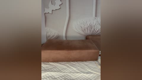 A video showing a brown leather ottoman with a white textured cushion on a beige quilted blanket. The background features a white wall with decorative star and wave designs. The scene exudes a cozy, minimalist aesthetic.
