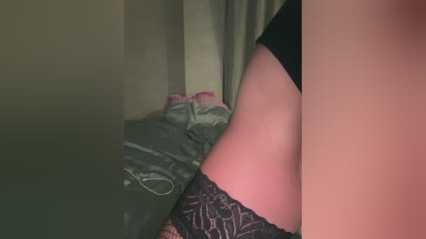 Video of a person's lower torso, showing a side view of a fair-skinned person with a slim build, wearing black lace panties. The background includes a bed with a gray comforter and a pink pillow, and a beige curtain.