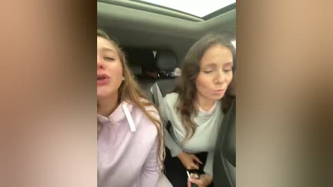 Video of two women in a car; one has long hair, light skin, and wears a light pink sweater, the other has shoulder-length dark hair, fair skin, and wears a gray sweater.