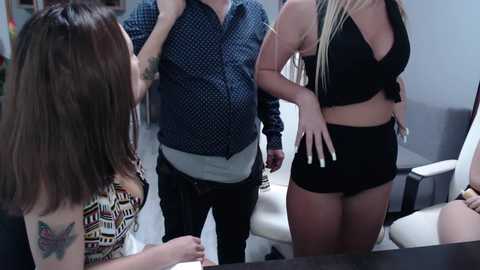 Video of three people in a modern office. A man in a blue shirt and black pants, a woman with long blonde braids in a black halter top and shorts, and a woman with brown hair in a patterned top.