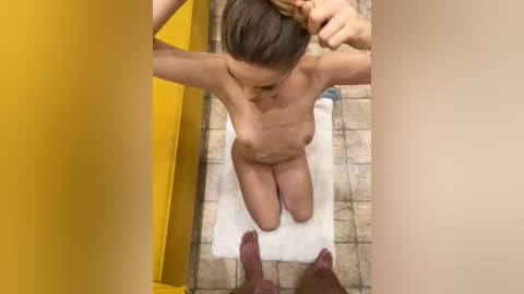 Video of a naked woman with small breasts, fair skin, and brown hair, kneeling on a white towel on tiled floor, seen from above, with legs spread, hands holding hair.