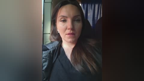 Video of a serious-looking woman with long brown hair, wearing a black dress, in a dimly lit room with a blue curtain in the background.