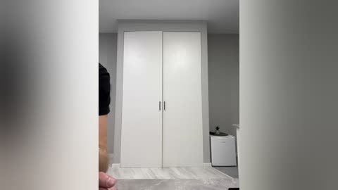 Video of a minimalist, modern walk-in closet with white cabinetry and drawers, featuring sleek silver handles. The room has light grey walls and a light wooden floor. A person in a black top is partially visible on the left.