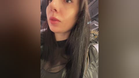 Video of a young woman with light skin and straight black hair, wearing a black top and a puffy, dark green jacket. She has a neutral expression, with slightly pouty lips. Background is blurred, suggesting an indoor setting.