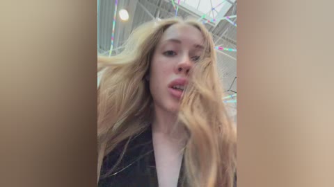 Video of a young, fair-skinned woman with long, blonde hair blowing in the wind. She's wearing a black jacket and has a slightly open mouth. The background features a modern indoor setting with colorful string lights.