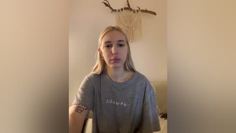 Video of a young, fair-skinned, blonde woman wearing a gray t-shirt with Greek text, standing in a minimalist room with beige walls, a macrame wall hanging, and a small tattoo on her arm.