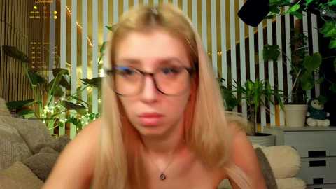 Video of a blonde woman with glasses, wearing a necklace, in a room with plants and a teddy bear, blurred background.