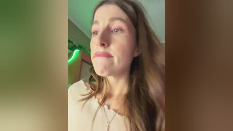 Video of a fair-skinned woman with long brown hair and pink lipstick, wearing a beige blouse, in a softly lit room with a green light.