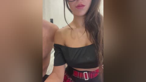 Video of a young woman with fair skin, long brown hair, wearing a black off-the-shoulder crop top, red plaid belt, and black high-waisted pants, standing in a narrow hallway.