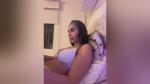 Video of a young, light-skinned woman with long brown hair, wearing a white bra, lying on a bed with white and heart-patterned pillows, in a dimly lit room.