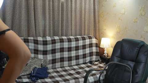 Video of a woman in a black bra and panties standing beside a plaid couch with a blue handbag on it, next to a dark leather office chair. The background features patterned wallpaper and a lit table lamp.