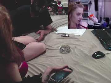 Video of a dimly lit bedroom with two young women, one with red hair and another with blonde hair, sitting on a beige bed, using smartphones. A close-up inset shows the blonde woman holding an orange object.