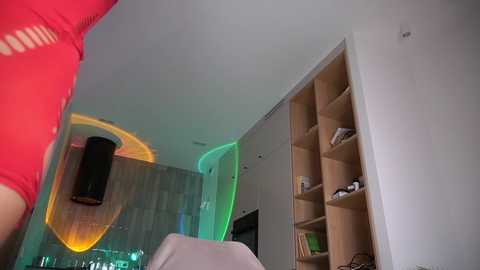 Video of a modern, well-lit room with a red shirted person, wooden bookshelves, and a wall-mounted TV, highlighted by ambient green and orange lighting.