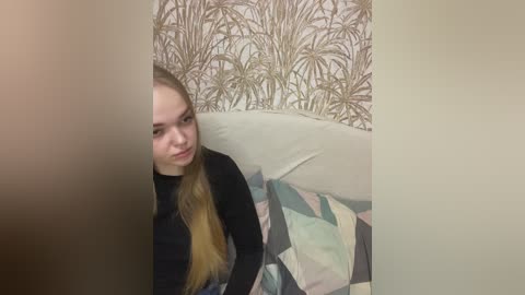Video of a young Caucasian woman with long blonde hair, wearing a black top, sitting on a sofa with a patterned cushion. The background features a beige wall with a botanical wallpaper.