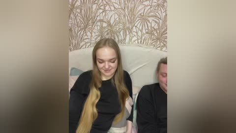 Video of a young blonde woman with long hair, wearing a black sweater, sitting on a beige couch, surrounded by a beige and gold-patterned wallpaper.