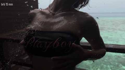 Video of a wet, topless woman with wet, shoulder-length hair, holding a black Playmate magazine. She stands by a wooden railing, overlooking a turquoise ocean. Water droplets cover her skin, creating a glistening effect.