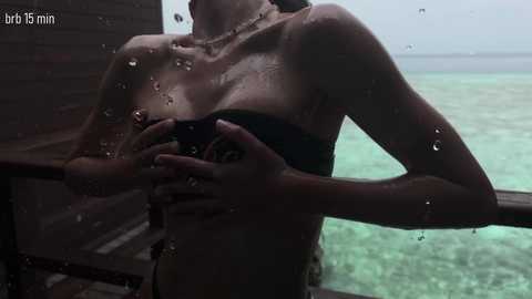 Video of a wet, topless woman with light skin, medium breasts, and wet hair, standing on a wooden deck overlooking turquoise ocean water.