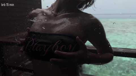 Video of a topless woman with wet, dark hair, holding a Playboy magazine against her breasts, standing on a wooden deck by a turquoise ocean.