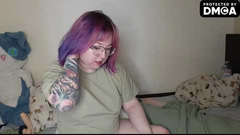 Video of a pale-skinned woman with purple hair, wearing glasses, green shirt, and colorful arm tattoo, sitting on a bed with patterned bedding, in a dimly-lit room.