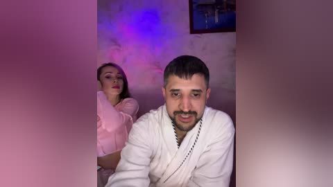 Video of a man in a white robe with a mustache, looking at a woman in a pink top, both partially obscured by a pink and purple haze, in a dimly lit room with a painting on the wall.