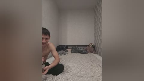 A video shows a shirtless, slim Asian man with short dark hair kneeling on a bed in a dimly lit, sparsely furnished room with beige walls and patterned wallpaper.