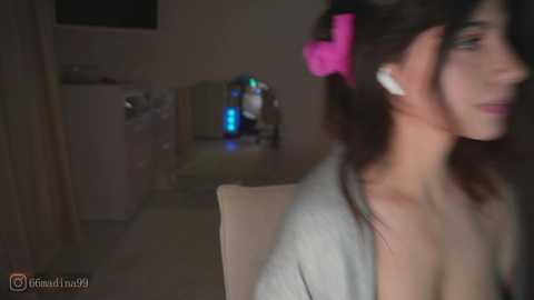 A dimly-lit video shows a young woman with fair skin, wearing a pink hair bow and gray cardigan, sitting in a chair, her profile facing left. The background is a blurred room with a TV, blue light, and a trash can.