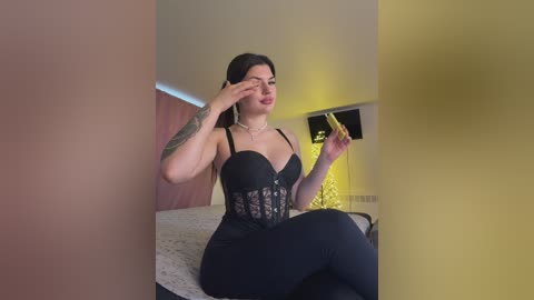 A video of a curvy woman with long black hair, wearing a black lace-up corset and tight black pants, sitting on a bed. She has a tattoo on her left arm.