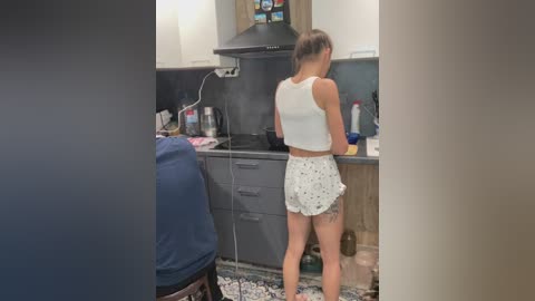 Video of a young woman with short brown hair, wearing a white crop top and floral shorts, cooking in a modern kitchen with white cabinets, black backsplash, and a stove.
